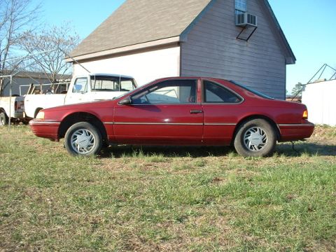 1989 Ford Thunderbird - A red 1989 Ford Thunderbird coupe is parked on a grassy lot next to a white truck and a building.