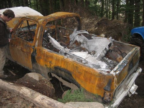 1990 Ford Bronco - A severely damaged pickup truck sits in a wooded area, with another vehicle partially visible.