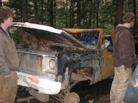 1990 Ford Bronco - This severely damaged pickup truck sits in a wooded area with two onlookers.