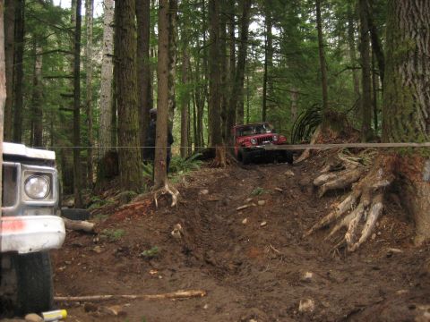 1990 Ford Bronco - Two off-road vehicles navigate a challenging muddy trail through a dense forest.