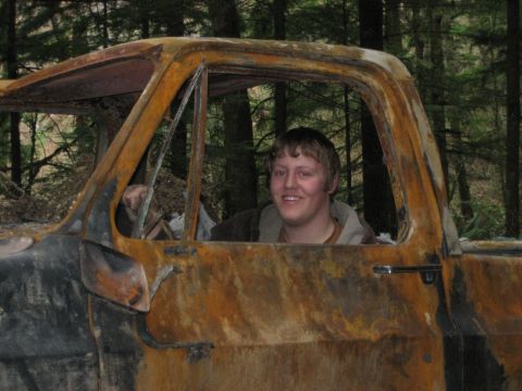 1990 Ford Bronco - A person sits in the driver's seat of a heavily damaged and burnt truck cab, surrounded by trees.