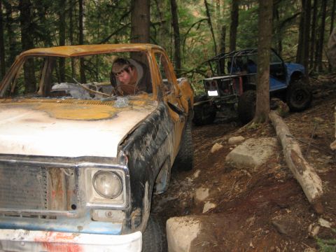1990 Ford Bronco - Two off-road vehicles, one a heavily damaged pickup, are parked on a wooded trail.