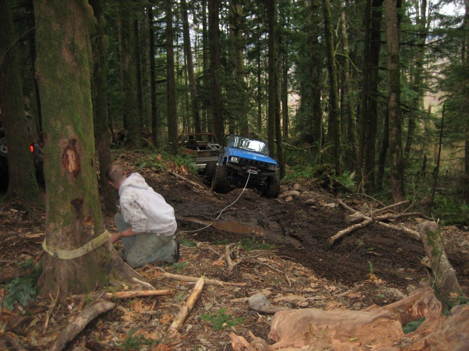 Two off-road vehicles are stuck in mud on a forest trail, one blue SUV being pulled by a winch attached to a tree