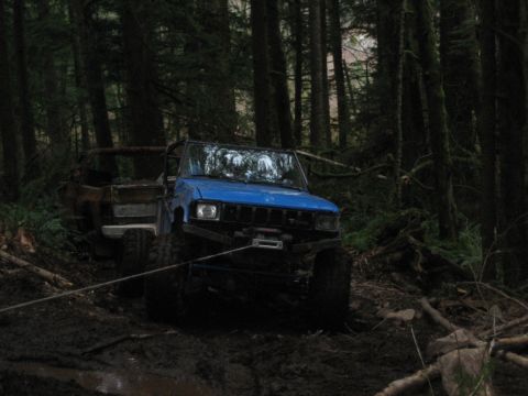 1990 Ford Bronco - Two vehicles are stuck in the mud in a dense forest, with one vehicle being pulled by a tow rope.