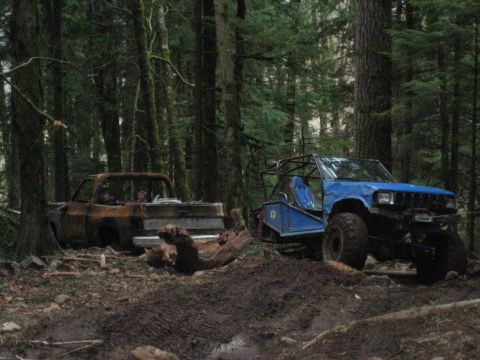 1990 Ford Bronco - Two modified off-road vehicles sit in a wooded area, one appearing burnt and the other a blue buggy.
