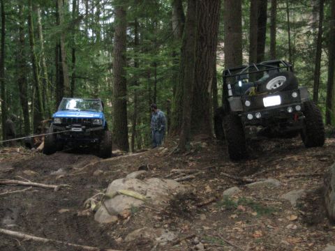 1990 Ford Bronco - Two off-road vehicles navigate a muddy, wooded trail with one vehicle appearing to be winched.