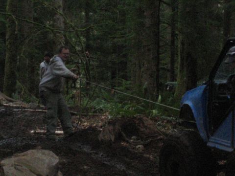 1990 Ford Bronco - Two men use a rope to pull a blue SUV out of the mud in a forest setting.