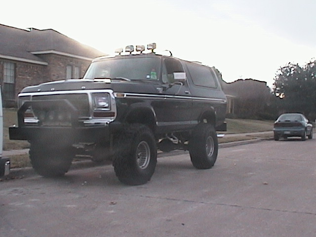Black lifted Ford Bronco backlit by sun in a residential street, roof lights and brush bar visible