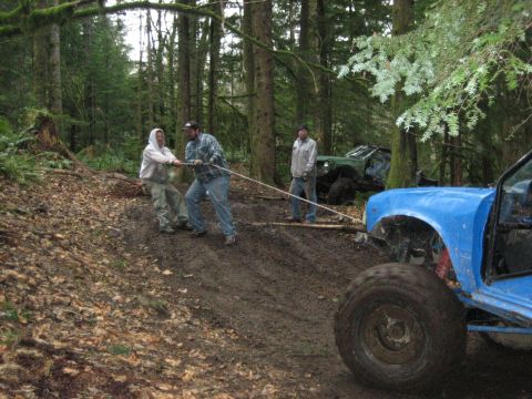 1990 Ford Bronco - Two men pull a rope to help a blue off-road vehicle navigate a muddy trail in a wooded area.