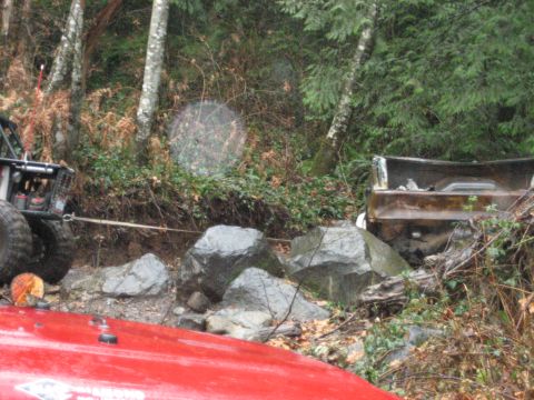 1990 Ford Bronco - An off-road vehicle tows a stuck pickup truck out of a rocky, wooded area.