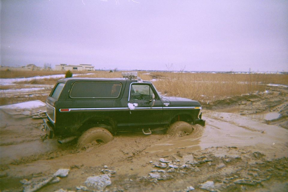 Black Ford Bronco sunk into a muddy rut in a snow-dusted field, side rear view
