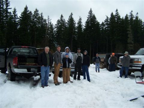 1996 Ford Bronco - Group of nine people in winter coats and hoodies stand in deep snow at a wheeling meet-up, backed by a dark evergreen forest under an overc…