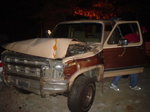 1984 Ford Bronco - This 1980s SUV has significant front-end damage, including a crumpled hood and bent bumper, with the driver's door ajar.