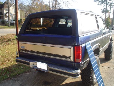 1984 Ford Bronco - The rear of a blue Ford Bronco is shown with a chrome bumper and gold trim panel.