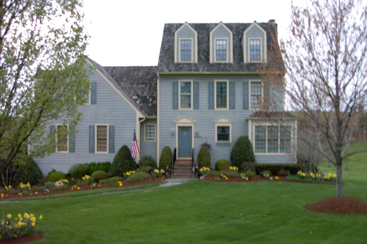 Two-story gray colonial house with lawn, flower beds, and an American flag.