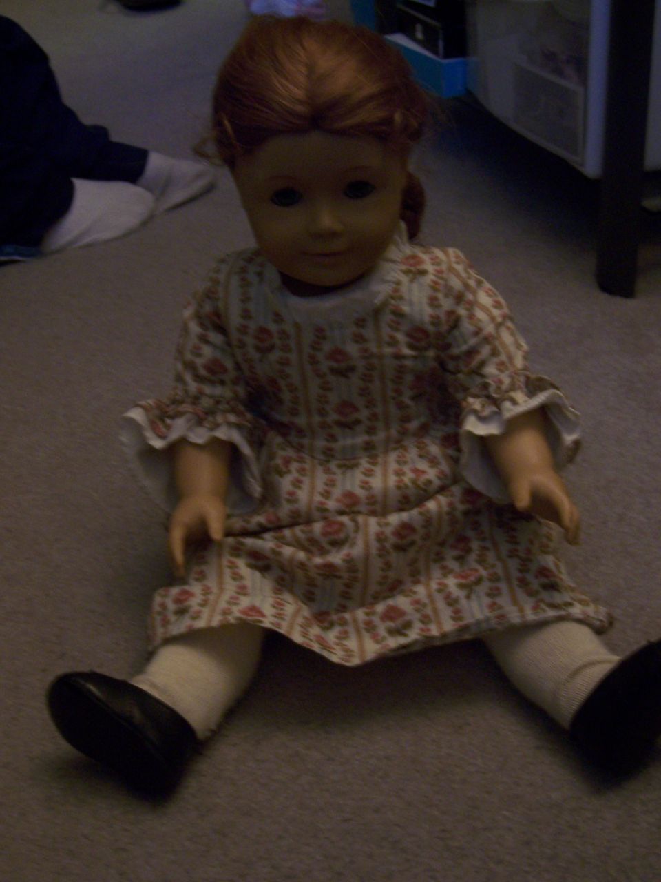 Doll with red hair wearing a floral dress and white socks with black shoes, sitting on carpet