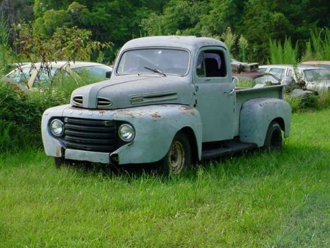 1972 Ford Ranchero - This classic Ford pickup truck, likely from the late 1940s, sits in a field surrounded by overgrown vegetation and other abandoned vehicles.