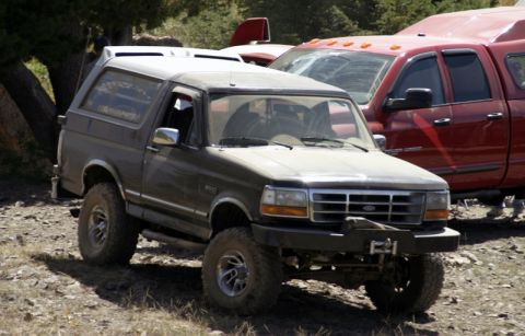 1991 Ford Bronco - A lifted Ford Bronco sits parked on a dirt lot next to a red pickup truck.