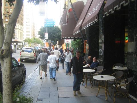 1990 Ford Bronco - Pedestrians walk along a downtown city sidewalk past a cafe with outdoor tables and parked cars at the curb.
