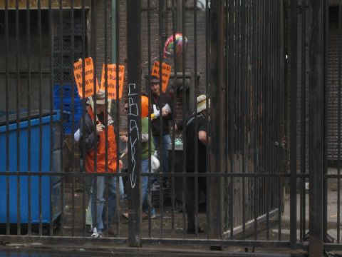 1990 Ford Bronco - Demonstrators in orange vests hold picket signs and a drum behind a black metal fence in a city alley.