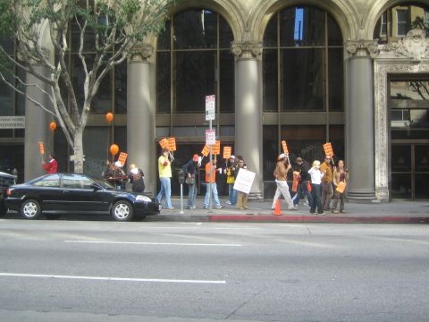1990 Ford Bronco - Picketers carrying orange signs and balloons march along a city sidewalk past a parked black car and stone columns.