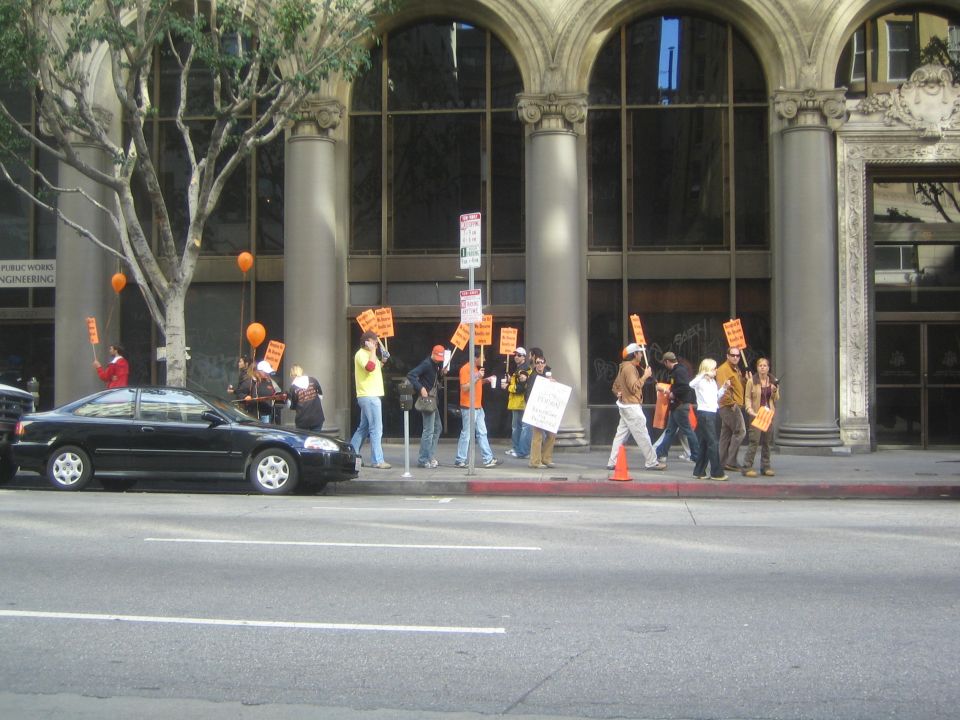 Picketers with orange signs and balloons marching on a sidewalk past a black car