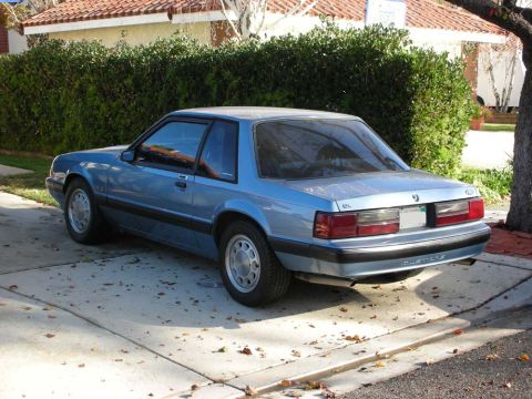 1990 Ford Mustang - A blue coupe is parked on a concrete driveway with fallen leaves scattered around.