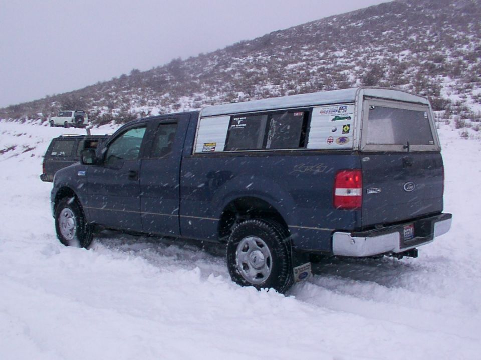 Blue Ford F-150 truck with a white camper shell driving in snow, another truck behind it