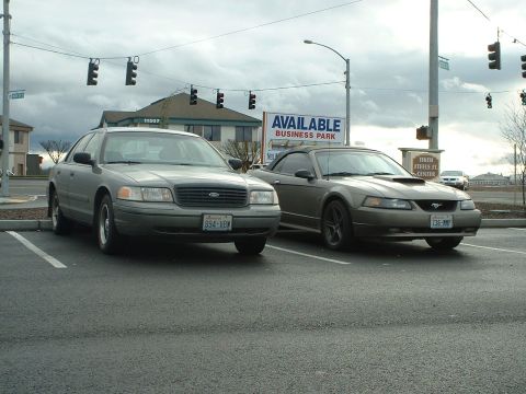 2000 Ford Crown Victoria - A tan Ford Mustang convertible is parked next to a gray sedan under an overcast sky.