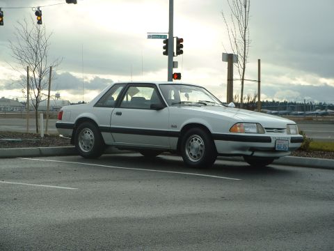 2000 Ford Crown Victoria - This white 1987 Ford Mustang coupe is parked in a lot under an overcast sky.