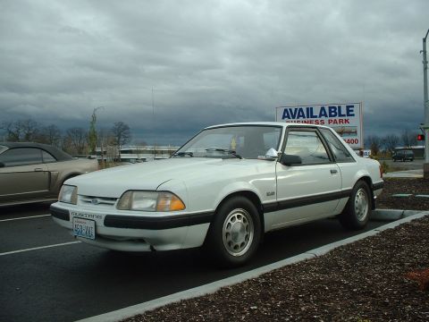2000 Ford Crown Victoria - A white 1987 Ford Mustang is parked in a lot next to another Mustang under a cloudy sky.