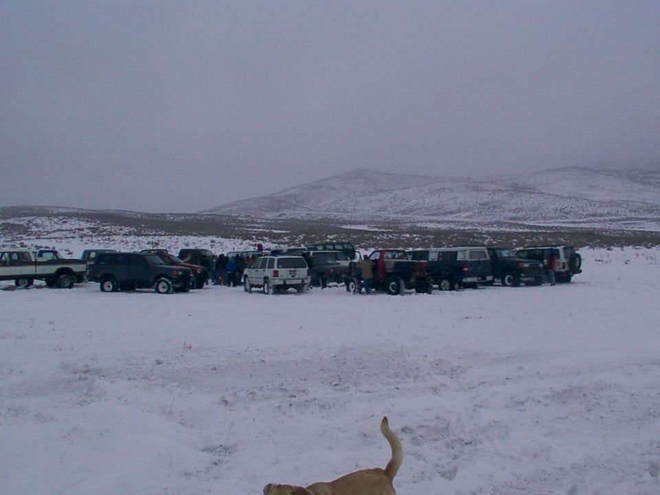 Several SUVs and pickup trucks parked in a snow-covered field with a group of people standing around