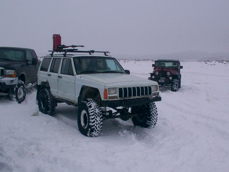 white Jeep Cherokee XJ with roof rack and red gas can, next to a black truck and a red Jeep Wrangler in the snow