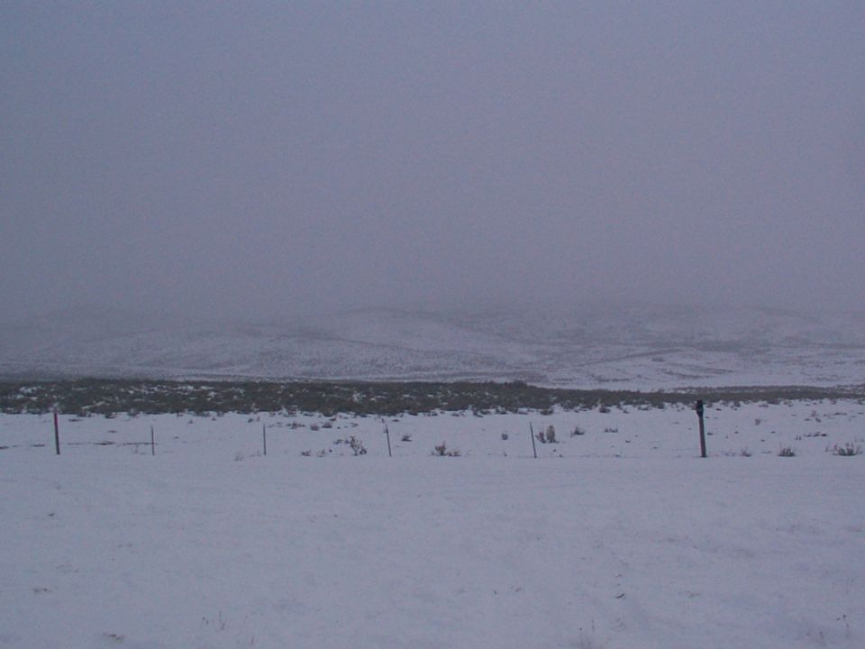 Snowy landscape with fence posts and distant hills under a hazy sky