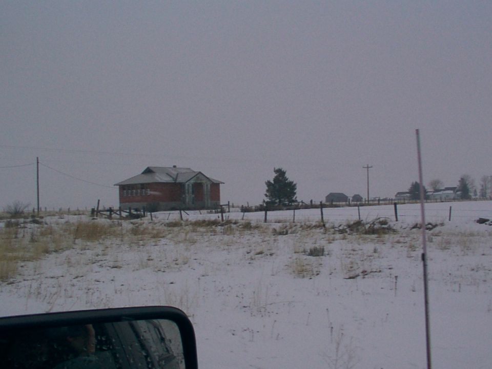 Snowy field with a brick building and fence, viewed from a vehicle