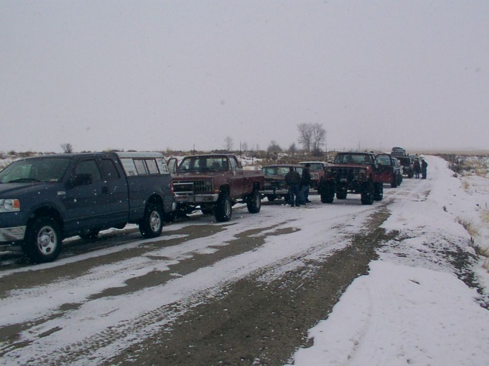 Several trucks lined up on a snowy road. The first truck is dark blue with a cap. Others are maroon and appear to be older models