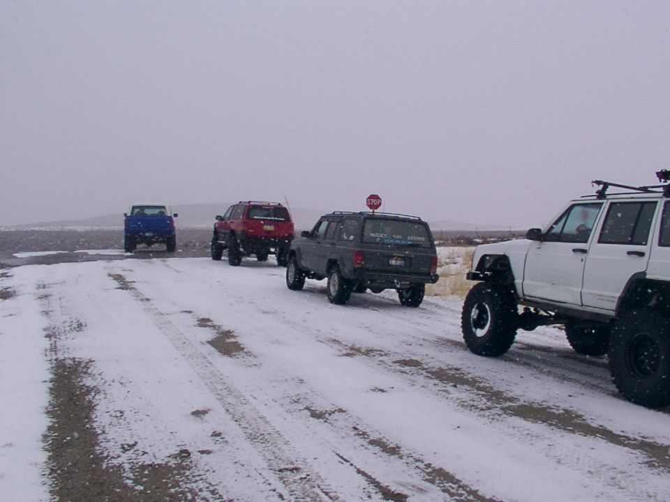 Four modified SUVs drive on a snow-covered dirt road under a cloudy sky