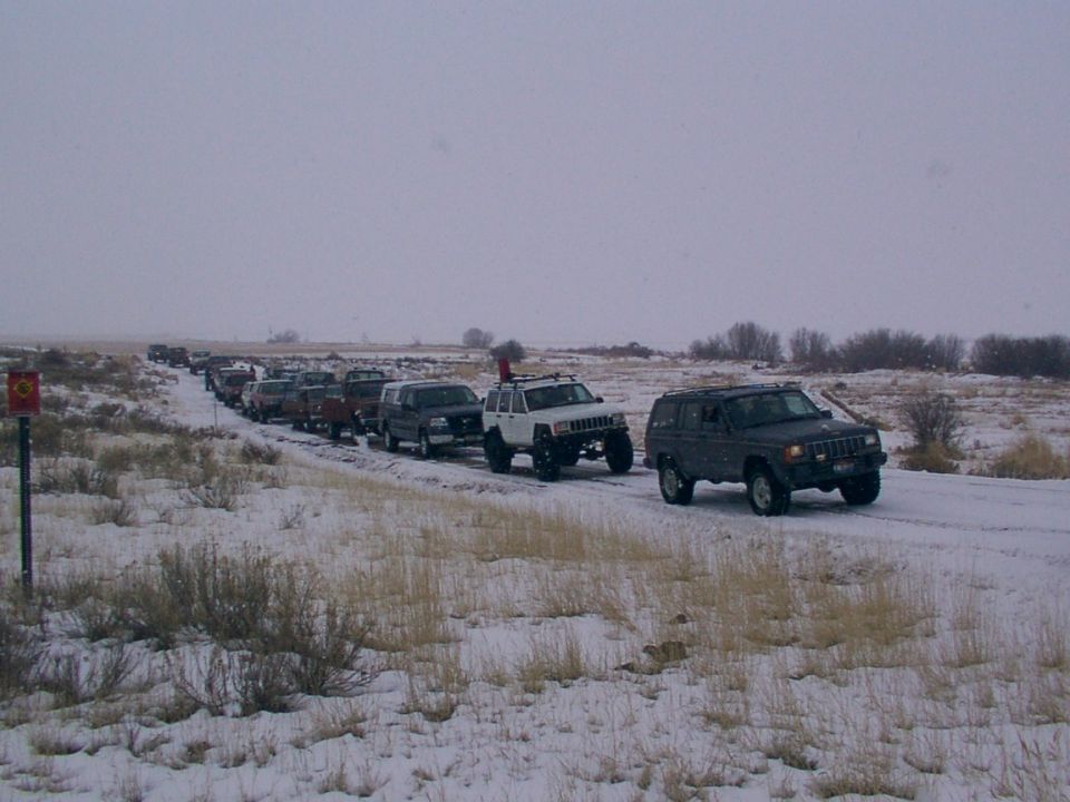 Line of SUVs and trucks on a snowy road, one white Jeep Cherokee with roof rack in foreground