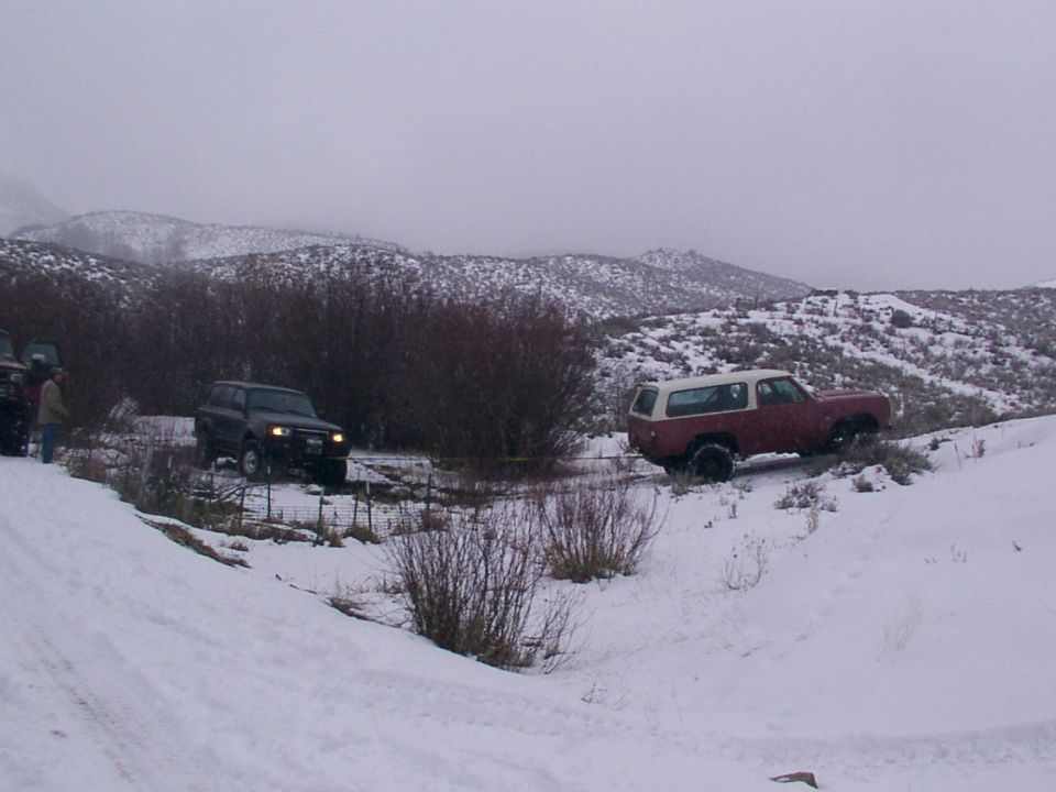Red and white SUV with a white camper shell on a snowy hill, with a dark SUV on the left