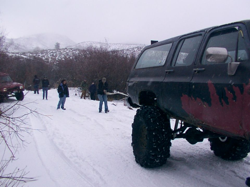 Off-road SUV with large tires in snow, people standing around it. Another red vehicle visible on left
