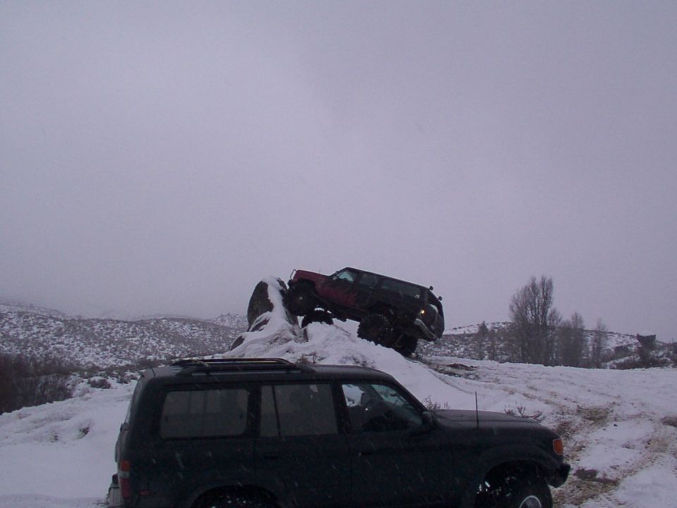 Two SUVs off-roading on a snowy hill; one is dark colored in the foreground, the other is red and climbing a rock