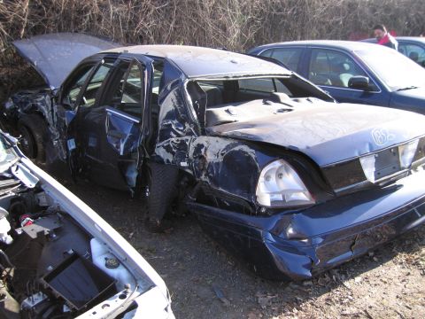 1991 Ford LTD Crown Victoria - This severely damaged sedan sits in a junkyard, with its hood open and rear end crushed.