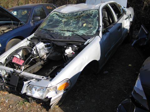 1991 Ford LTD Crown Victoria - A damaged silver sedan sits in a junkyard, its windshield shattered and hood removed.