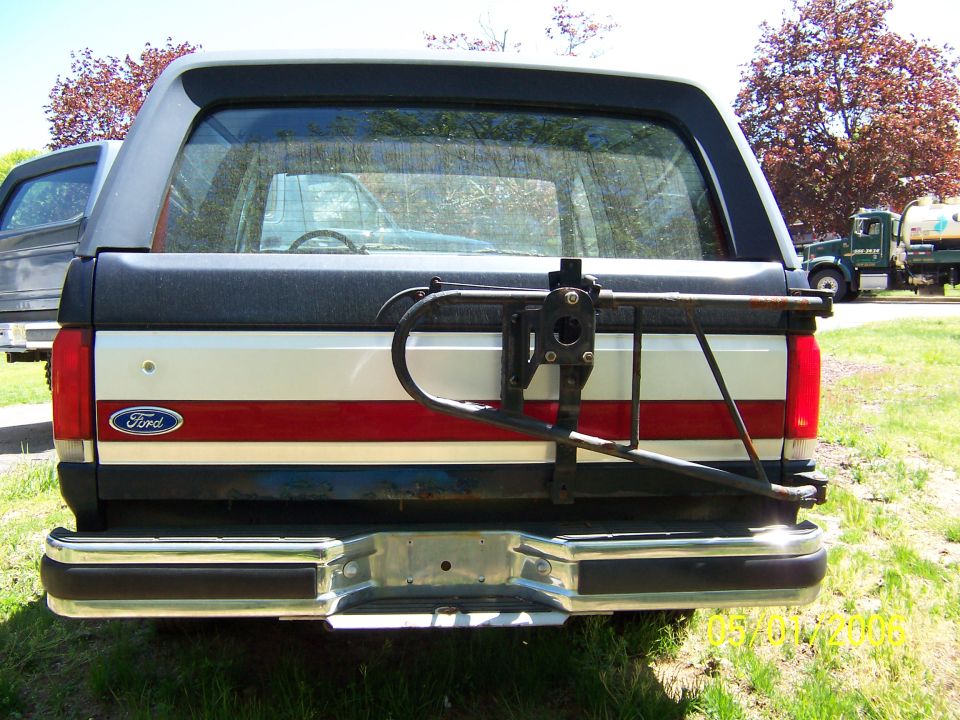 1987 Ford Bronco rear bumper, taillights, and swing-away spare tire carrier. Another vehicle is visible to the left