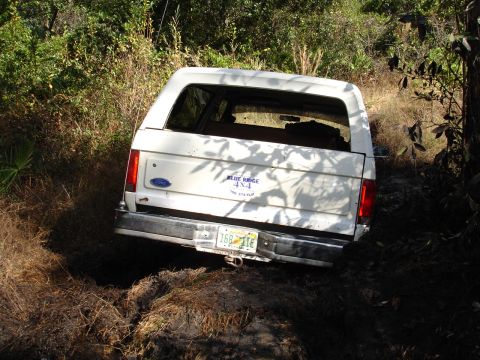 1989 Ford Bronco - A white Ford Bronco is stuck in a muddy, wooded area, with its rear end facing the camera.