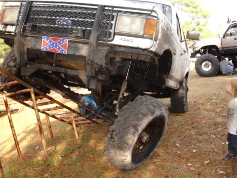 1989 Ford Bronco - A heavily modified truck with large tires is positioned on a metal ramp, with another lifted truck visible in the background.