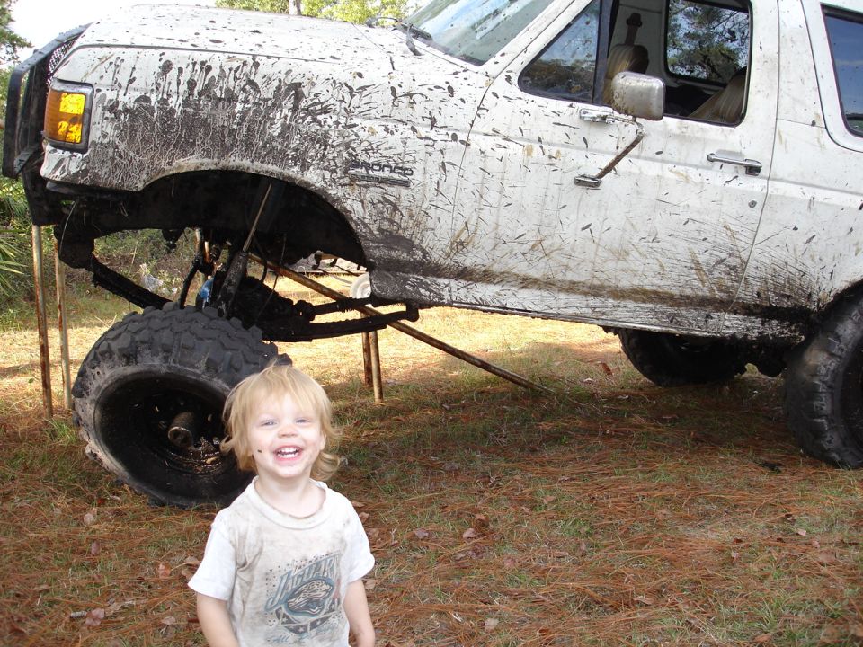 Mud-splattered white SUV, lifted with large tires, seen from the front-right. A smiling child stands in front