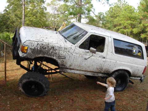 1989 Ford Bronco - Mud-covered lifted SUV, likely a Ford Bronco, with a child pointing. Lift kit and large tires are prominent.
