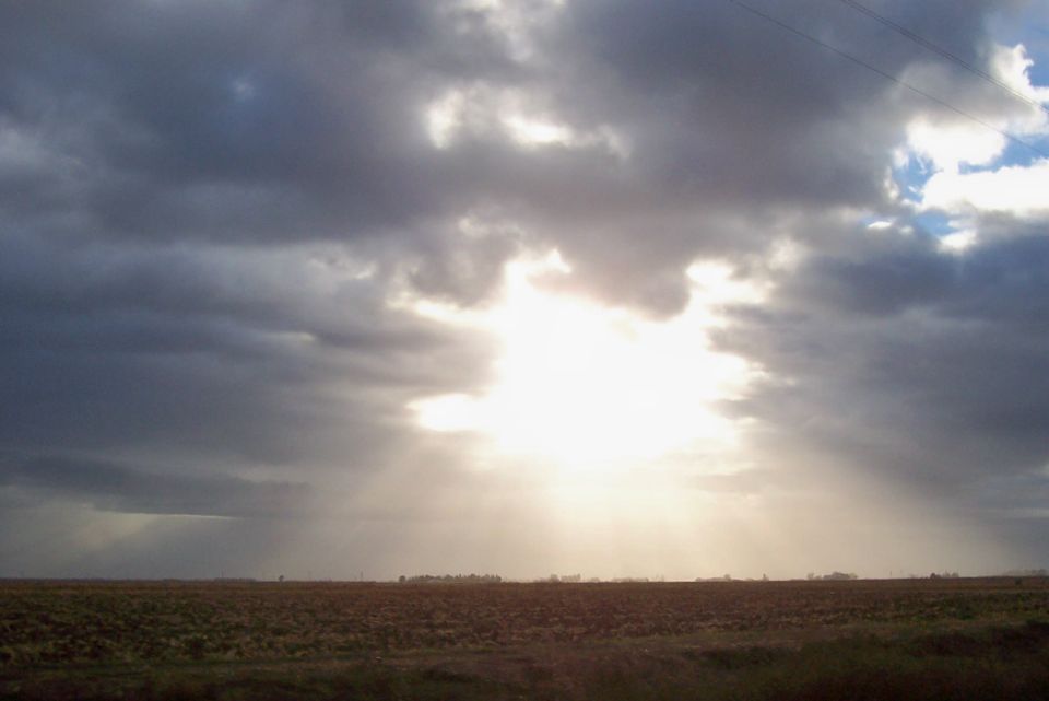 Overcast sky with sunbeams breaking through, over a flat, brown field