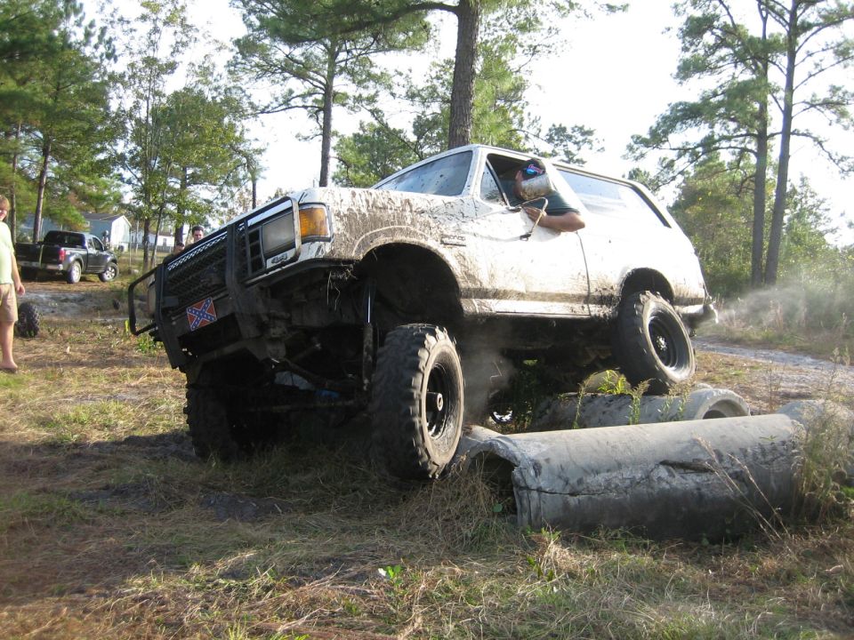 Muddy SUV climbing over concrete pipes, driver visible. Another truck in background